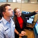 Two individuals are working together at a multi-monitor control or operations center workstation. A uniformed male officer and a woman in business attire are reviewing data displayed on screens, with the woman pointing at one of the monitors. The setting appears to be a professional command, security, or operations control room.