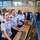 A group of military cadets or officers in light blue uniforms are gathered around computer workstations in what appears to be a modern operations or control room. They are attentively looking at large monitors, with one female cadet prominently positioned at the keyboard. Several civilians in casual business attire are also present in the background, suggesting a demonstration or tour.
