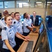 A group of military cadets or officers in light blue uniforms are gathered around computer workstations in what appears to be a modern operations or control room. They are attentively looking at large monitors, with one female cadet prominently positioned at the keyboard. Several civilians in casual business attire are also present in the background, suggesting a demonstration or tour.