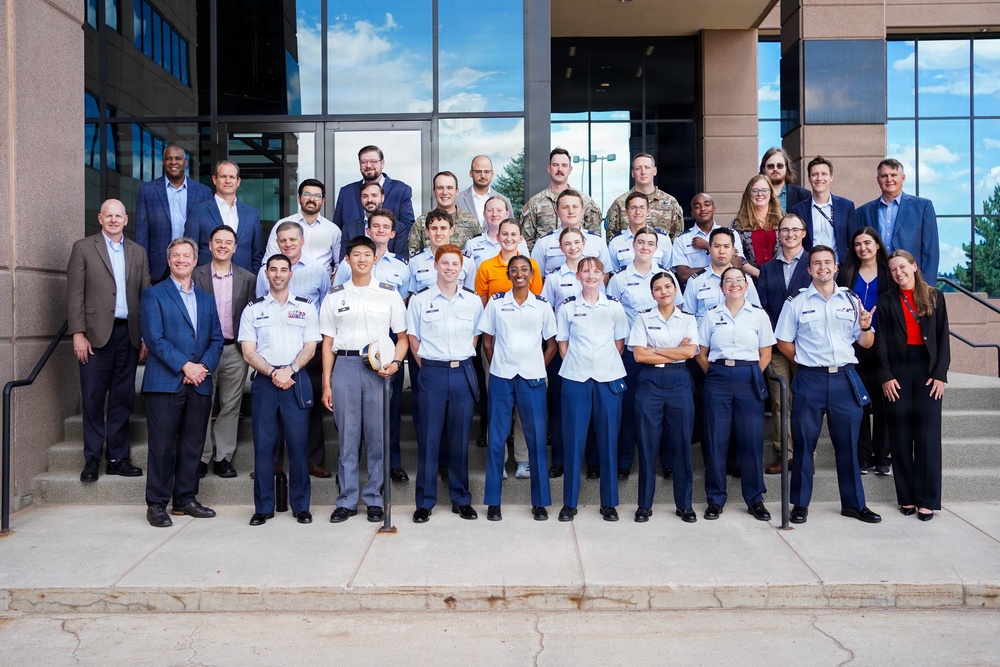 A large group photo taken on the steps of a modern institutional building, featuring approximately 30-35 individuals. The group includes military personnel in uniform (light blue shirts with dark blue trousers, consistent with U.S. Air Force or Space Force dress uniforms), civilians in business attire, and at least two individuals in military camouflage uniforms. The photo appears to be a formal group portrait, likely commemorating a program, event, or visit.