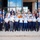 A large group photo taken on the steps of a modern institutional building, featuring approximately 30-35 individuals. The group includes military personnel in uniform (light blue shirts with dark blue trousers, consistent with U.S. Air Force or Space Force dress uniforms), civilians in business attire, and at least two individuals in military camouflage uniforms. The photo appears to be a formal group portrait, likely commemorating a program, event, or visit.