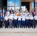 A large group photo taken on the steps of a modern institutional building, featuring approximately 30-35 individuals. The group includes military personnel in uniform (light blue shirts with dark blue trousers, consistent with U.S. Air Force or Space Force dress uniforms), civilians in business attire, and at least two individuals in military camouflage uniforms. The photo appears to be a formal group portrait, likely commemorating a program, event, or visit.
