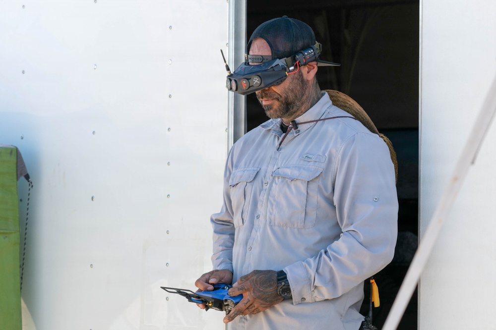 A bearded man wearing a baseball cap with FPV (First Person View) goggles mounted on his head stands near a white trailer or vehicle. He is holding what appears to be a drone remote controller in his hands, suggesting he is operating or preparing to operate an FPV drone. The man has visible tattoos on his hands and is wearing a light gray long-sleeve work shirt.