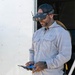 A bearded man wearing a baseball cap with FPV (First Person View) goggles mounted on his head stands near a white trailer or vehicle. He is holding what appears to be a drone remote controller in his hands, suggesting he is operating or preparing to operate an FPV drone. The man has visible tattoos on his hands and is wearing a light gray long-sleeve work shirt.