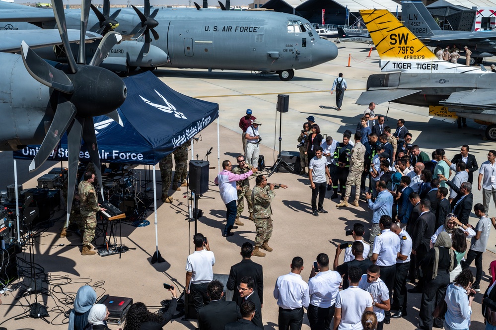 A United States Air Force outdoor event or airshow reception is taking place on an aircraft ramp/tarmac. Military personnel and civilians are gathered around a live band performing under a U.S. Air Force branded tent, with multiple military aircraft visible in the background. The event appears to be a public engagement or international relations function given the diverse crowd in both military and civilian attire.