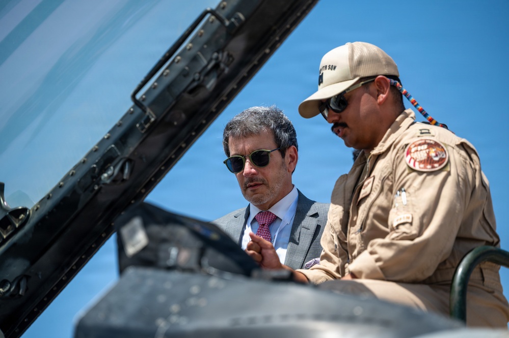 Two individuals are examining the cockpit area of a military aircraft. One person is wearing a gray business suit with a pink tie and sunglasses, while the other is dressed in a tan/khaki flight suit with patches and a matching cap. They appear to be engaged in a briefing or demonstration near the aircraft.