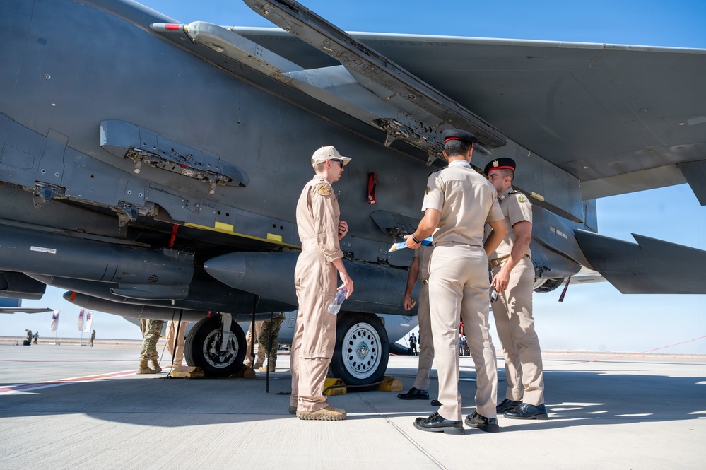 Military personnel from what appear to be two different nations are conducting a close inspection of a large military aircraft, likely a B-1B Lancer bomber, on a flight line or tarmac. One individual in a tan flight suit with a baseball cap appears to be briefing or explaining aircraft features to several others wearing tan dress uniforms with red shoulder boards. The setting suggests a military air show, exchange visit, or bilateral exercise.