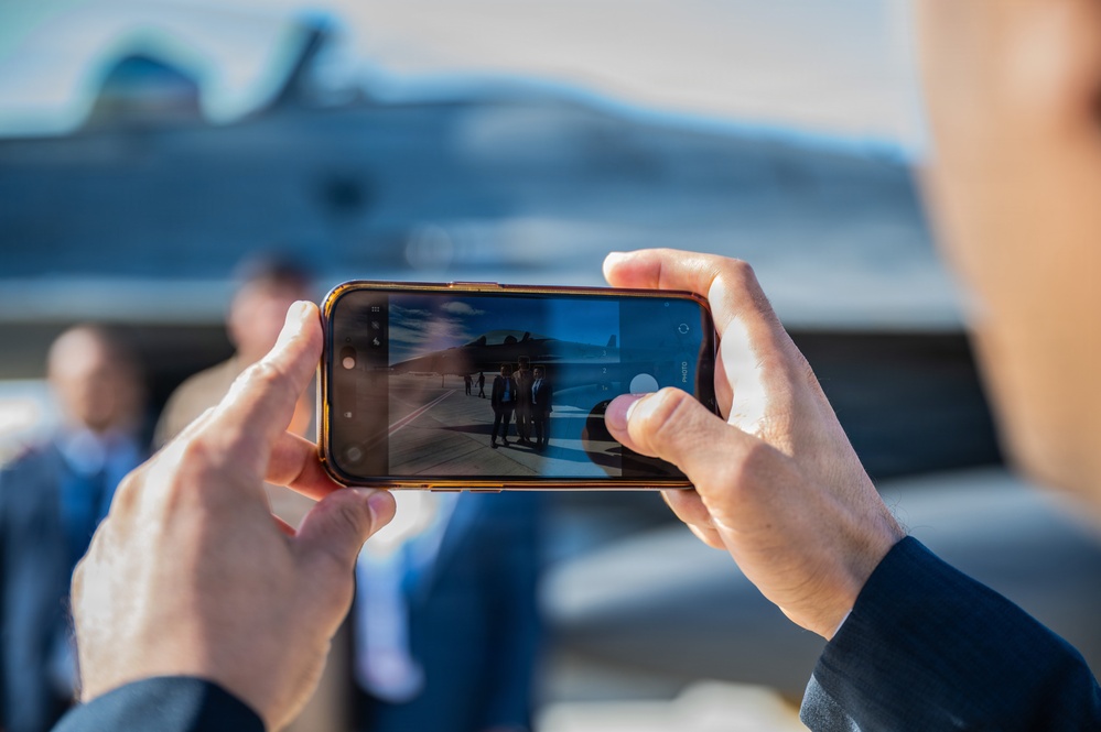 A person is photographing a group of individuals standing near a military jet aircraft using a smartphone held in landscape orientation. The phone screen displays the scene being captured, showing several people posed in front of what appears to be a fighter aircraft on a tarmac. The image uses a shallow depth of field, keeping the phone and screen in sharp focus while the background remains blurred.