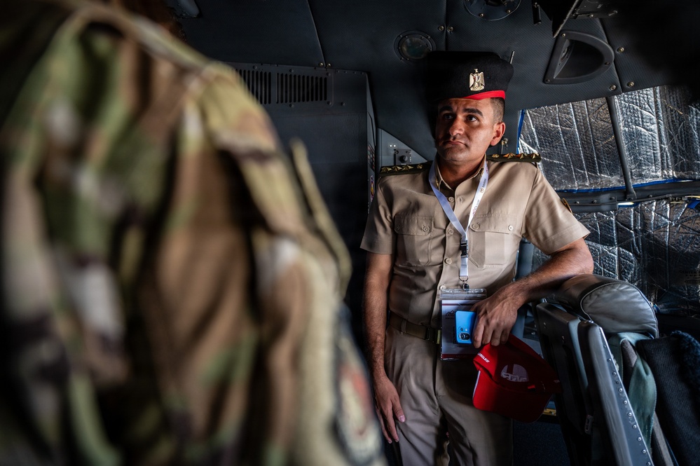 A military officer in a tan/khaki uniform with a black peaked cap bearing an eagle insignia sits inside what appears to be a military helicopter or transport aircraft. He is wearing a lanyard with an ID badge and holding a red cap, looking upward or to the side. In the foreground, an out-of-focus figure in camouflage gear is partially visible.