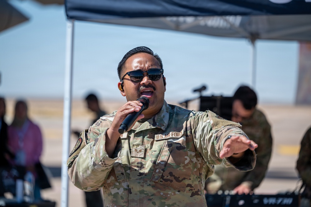 A U.S. Air Force service member is performing vocally at an outdoor military event, holding a microphone and gesturing with an outstretched hand. The individual is wearing OCP (Operational Camouflage Pattern) uniform with 'U.S. AIR FORCE' tape visible on the chest, along with sunglasses and an in-ear monitor. The setting appears to be on or near a flight line or airfield, likely during an air show or base event.
