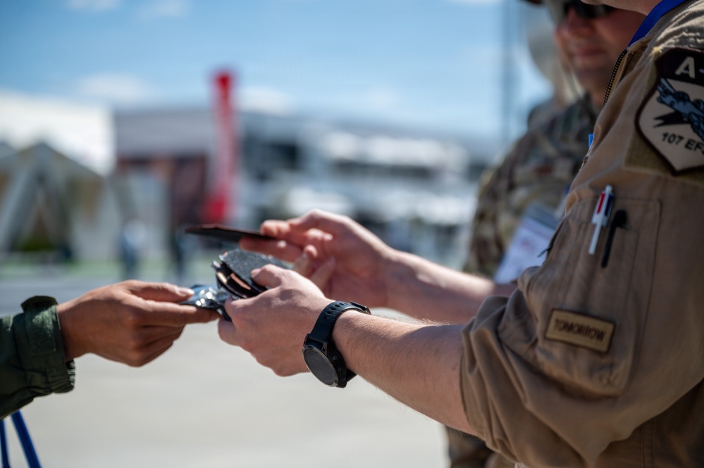 A military personnel member wearing a tan/brown flight suit with a '107 EF' squadron patch and a name tape reading 'TOMORROW' is exchanging or inspecting documents/credentials with another individual in an olive green flight suit. The interaction appears to be a credential check or document exchange at an outdoor location, likely an airshow or military event. The background shows blurred structures and a red banner, suggesting a public or semi-public venue.