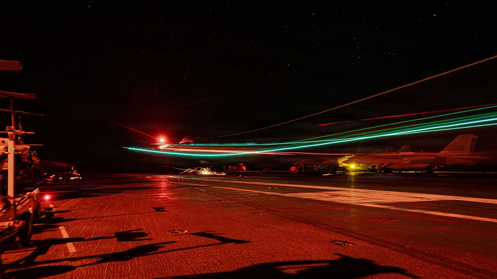 A long-exposure night photograph taken on an aircraft carrier flight deck, capturing light trails from aircraft navigation lights (red and green/teal) as a jet launches or taxis. The wet deck surface reflects the ambient red lighting used for night operations. At least one stationary aircraft is visible on the right side of the frame, with deck equipment and superstructure visible on the left.