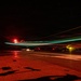 A long-exposure night photograph taken on an aircraft carrier flight deck, capturing light trails from aircraft navigation lights (red and green/teal) as a jet launches or taxis. The wet deck surface reflects the ambient red lighting used for night operations. At least one stationary aircraft is visible on the right side of the frame, with deck equipment and superstructure visible on the left.
