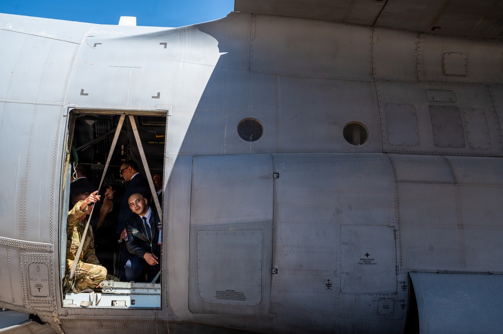Several individuals, including at least one person in military camouflage uniform and others in civilian or formal attire, are visible inside the open side door of a large military transport aircraft. The aircraft's fuselage dominates the frame, showing riveted aluminum panels, two circular porthole windows, and various access panels with stenciled markings. The scene appears to be a tour or inspection of the aircraft, likely at an airshow or military demonstration event.