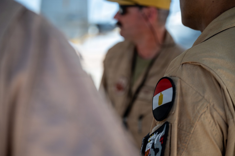 A close-up photograph focusing on the shoulder of a person wearing a tan/khaki military uniform with a circular Egyptian flag patch on the upper sleeve. A second individual wearing a yellow hard hat and tan uniform is visible in the background, slightly out of focus. The image appears to document a military or official interaction, likely in an outdoor setting.