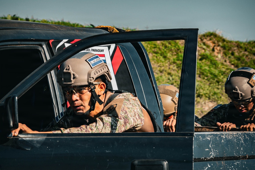 The image shows military personnel riding in tactical vehicles, wearing combat helmets and camouflage uniforms. At least two nations are identifiable from helmet patches: El Salvador (visible on the foreground soldier's helmet) and what appears to be Paraguay (text visible on a flag/banner inside the vehicle). The scene appears to be a military exercise or convoy operation.