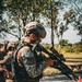 The image shows armed military personnel from El Salvador in full tactical gear during what appears to be a training exercise or field operation. The lead soldier is holding a rifle equipped with a scope and is wearing digital camouflage, a tactical vest, and a tan ballistic helmet. Additional soldiers are visible in the background, similarly equipped.