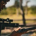 A person wearing digital camouflage clothing and a cap is holding and inspecting a revolver-style handgun in an outdoor setting. In the background, a scoped rifle is visible resting on what appears to be a shooting bench or rest. The setting appears to be an outdoor shooting range during daylight hours.