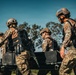 A group of military personnel in camouflage uniforms and tactical gear are carrying what appears to be a stretcher or equipment platform together. The soldiers are wearing ballistic helmets, body armor, and are armed, suggesting a training exercise or operational activity. The setting appears to be an outdoor field environment with trees visible in the background.