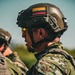 A Colombian military special forces soldier is photographed from behind and slightly to the side, wearing full tactical gear. The soldier displays a Colombian national flag patch on their helmet and a 'Fuerzas Comando' unit patch on their shoulder. A second soldier is partially visible in the background.
