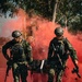 Three armed military personnel in camouflage uniforms are carrying a stretcher through a red smoke screen. The soldiers are equipped with tactical gear, helmets, and assault rifles, suggesting a combat casualty evacuation drill or training exercise. The red smoke grenade appears to be used as a visual screen or signal during the exercise.