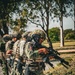 A group of military personnel in full tactical gear are lined up in a column formation, appearing to be conducting a training exercise or patrol drill. The lead soldier is visibly inspecting or handling a rifle, with a Mexican flag patch clearly visible on their shoulder. The setting is an outdoor environment with trees and open ground in the background.
