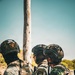 Three heavily equipped military or law enforcement personnel are gathered in a close huddle, appearing to confer or receive a briefing. They are wearing tactical gear including ballistic helmets with Mexican flag patches, plate carriers, and camouflage uniforms. The setting appears to be an outdoor, arid or semi-arid environment.