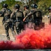 A group of five military or special operations personnel in full tactical gear are gathered together amid a red smoke grenade deployment in an open outdoor area. The personnel are wearing camouflage uniforms, ballistic helmets, and plate carrier vests, and appear to be conducting a training exercise or tactical drill. The red smoke creates a dramatic visual effect billowing across the ground in the foreground.