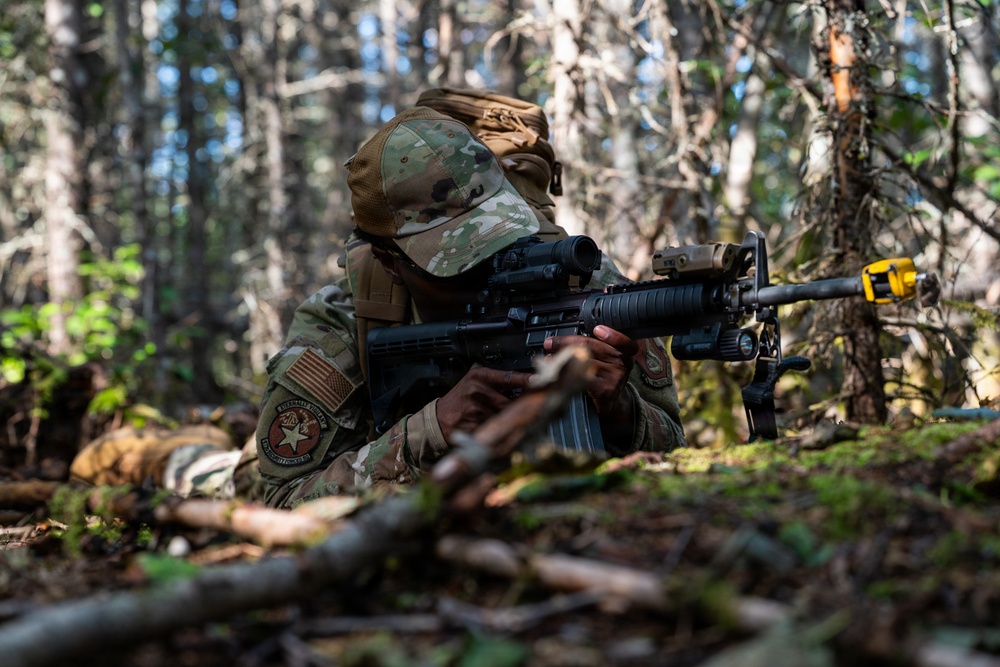 A soldier in MultiCam/OCP camouflage uniform is in a prone or low-crouch firing position in a forested environment, aiming an M4-style carbine with an optic and accessories. The individual wears a US flag patch and a unit patch on the left sleeve, along with a multicam baseball-style cap. The setting appears to be a training exercise based on the yellow training device visible on the weapon's muzzle.