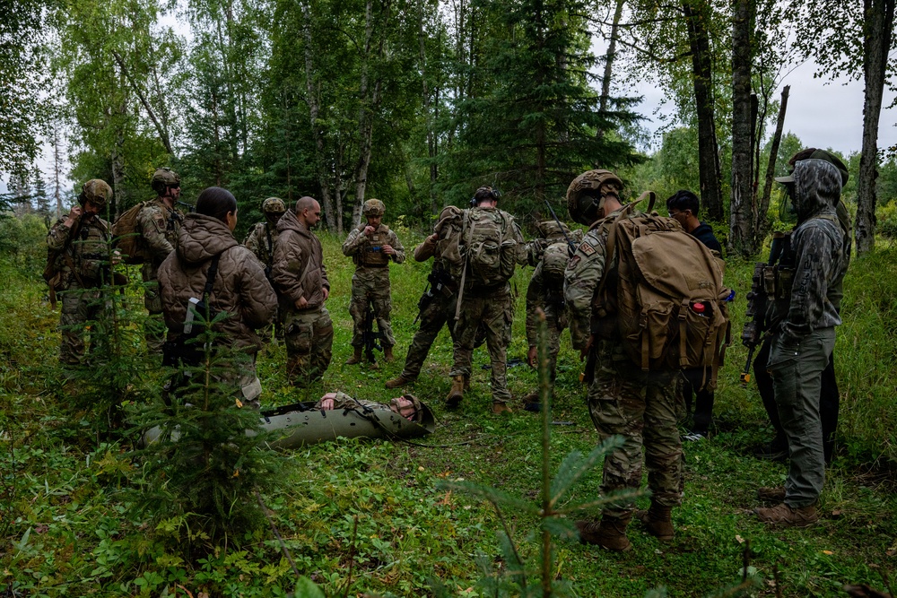 A group of military personnel in camouflage uniforms are gathered in a forested clearing, appearing to conduct a field training exercise. One individual is lying on the ground on what appears to be a litter or sled, suggesting a casualty evacuation or medical training scenario. The group includes personnel with tactical gear, backpacks, and weapons.