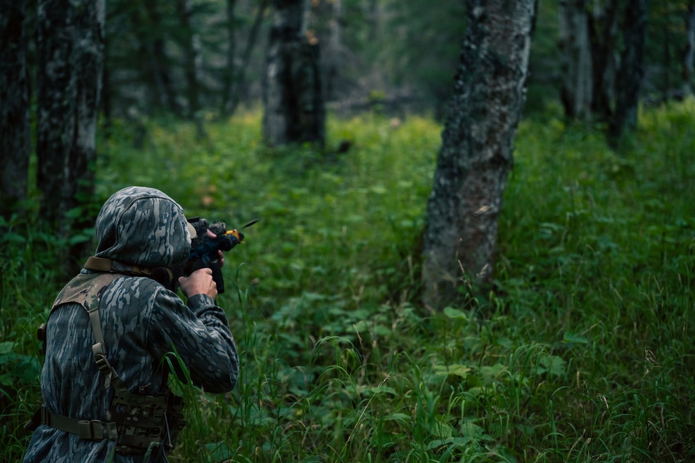 A person wearing full camouflage gear, including a hooded jacket, is aiming a rifle or airsoft weapon into a dense forest. The individual is positioned in the left portion of the frame, facing away from the camera into a green, tree-lined woodland environment. The scene appears to depict a military training exercise, hunting activity, or airsoft/paintball game.