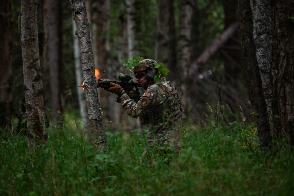 A U.S. Air Force Security Forces (SF) airman is photographed firing a weapon in a forested environment, with a visible muzzle flash captured at the moment of discharge. The soldier is wearing full multicam camouflage uniform, tactical vest, helmet with foliage, and hearing protection. The setting appears to be a training exercise in a dense woodland area.