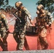 A group of military personnel in full combat gear are conducting what appears to be a tactical training exercise or operation. Several soldiers are carrying a stretcher or litter through a red smoke-filled environment, suggesting a casualty evacuation (CASEVAC) drill. The soldiers are wearing mixed camouflage patterns and are equipped with rifles, tactical vests, and helmets.