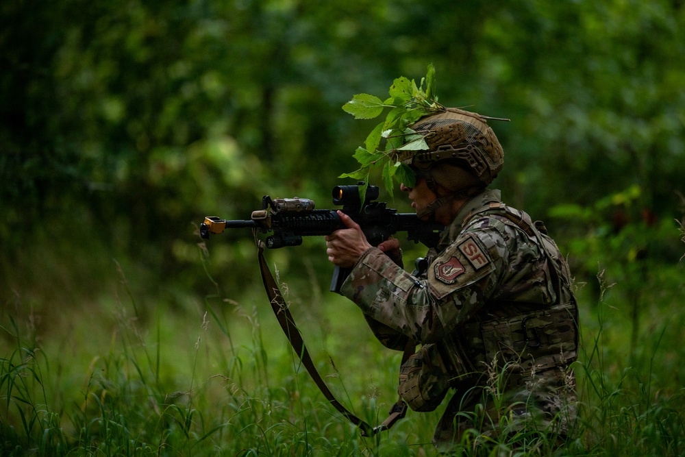 A soldier in full combat gear is positioned in a wooded, grassy area while aiming a rifle with a red dot optic sight. The individual is wearing multicam camouflage uniform with patches visible on the sleeve, a tactical helmet adorned with natural foliage for concealment, and a plate carrier vest. The setting appears to be a field training exercise or tactical operation in a lush green environment.