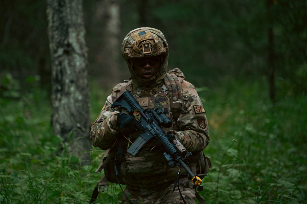 A U.S. Air Force Security Forces (SF) member is moving through a wooded area in full combat gear. The individual is wearing OCP (Operational Camouflage Pattern) uniform and carrying what appears to be an M4-style carbine with accessories. The setting suggests a field training exercise or operational patrol in a forested environment.
