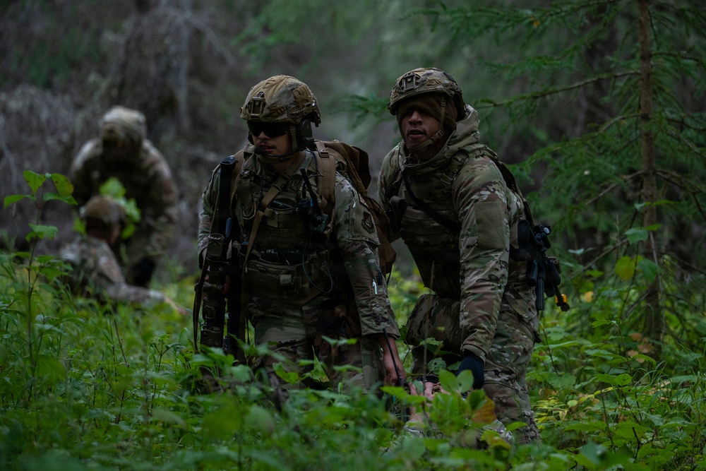 The image shows military personnel in full combat gear moving through a dense forested environment. Two soldiers are prominently visible in the foreground wearing Operational Camouflage Pattern (OCP) uniforms, tactical helmets, and body armor, with at least two additional personnel visible in the background. The scene appears to depict a field training exercise or patrol operation in a woodland setting.