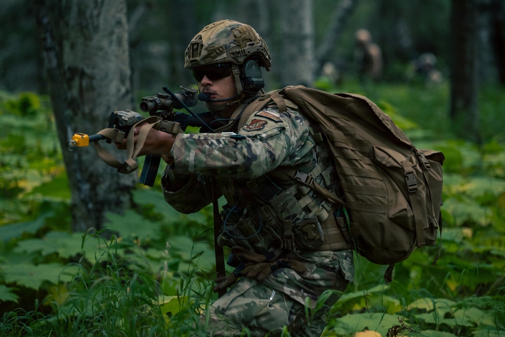 A military service member in full combat gear moves through a dense forest environment in a tactical posture. The soldier is equipped with a rifle fitted with accessories, a large rucksack, tactical vest, and ballistic helmet with communications equipment. Additional personnel are visible in the blurred background, suggesting a unit-level field training exercise or patrol operation.