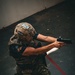 A person in full tactical gear is actively firing a semi-automatic pistol equipped with a red dot optic sight, captured mid-action at what appears to be an indoor shooting range or training facility. A spent brass casing is visibly ejecting from the firearm near the shooter's forearm. Multiple spent casings are scattered on the dark floor, indicating sustained firing activity.