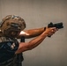 A tactical operator wearing multicam helmet and plate carrier is firing a semi-automatic pistol equipped with a red dot optic. A spent brass casing is visibly ejecting from the firearm, captured mid-air to the right of the weapon. The setting appears to be a training or operational environment against a plain concrete wall.