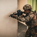 A soldier wearing multicam camouflage and a tactical helmet marked 'GRUMEC' is actively firing a rifle around a corner in what appears to be an urban combat or training environment. A spent brass casing is visibly mid-air to the left of the weapon, indicating the firearm has just been discharged. The soldier is equipped with full tactical gear including body armor, a backpack, and a weapon-mounted optic.
