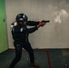 A tactical law enforcement or military officer wearing full black gear and a Dominican Republic flag patch is firing a handgun at a shooting range or training facility. The image captures the moment of discharge, with visible smoke/gas expelled from the muzzle. The setting appears to be an indoor training environment with a marked floor boundary.