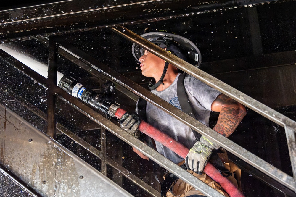 A firefighter is positioned on a metal staircase or ladder structure inside a smoke-darkened building, holding a red fire hose with a nozzle attachment. The individual is wearing protective gear including a helmet with face shield, tactical gloves, and has visible tattoos on both arms. Water droplets are visible throughout the scene, indicating active firefighting operations.