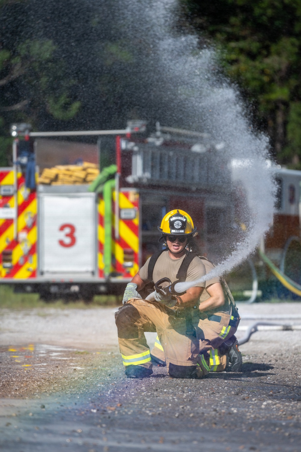 A firefighter in partial turnout gear is kneeling on the ground while operating a fire hose, directing a wide spray of water. A red fire engine marked with the number '3' and yellow chevron striping is visible in the background. The scene appears to be a training exercise or drill conducted outdoors.