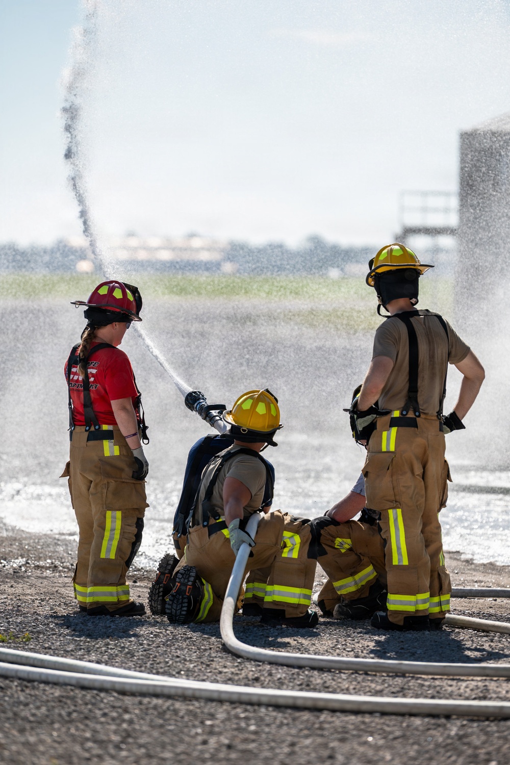 Three firefighters in full turnout gear are conducting a hose training exercise outdoors on a paved surface. One firefighter is kneeling and operating a fire hose nozzle, directing a high-pressure water stream upward, while two others stand nearby observing or assisting. The setting appears to be a training ground, likely at a fire academy or airfield facility.