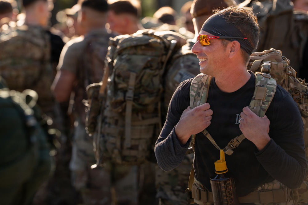 A smiling man in the foreground wears a black long-sleeve shirt, a dark headband, orange-tinted sport sunglasses, and a camouflage tactical backpack with a water bottle attached. He is surrounded by a group of similarly equipped individuals in a military or paramilitary training context. The image is captured in warm, low-angle light suggesting early morning or late afternoon.