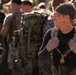 A smiling man in the foreground wears a black long-sleeve shirt, a dark headband, orange-tinted sport sunglasses, and a camouflage tactical backpack with a water bottle attached. He is surrounded by a group of similarly equipped individuals in a military or paramilitary training context. The image is captured in warm, low-angle light suggesting early morning or late afternoon.