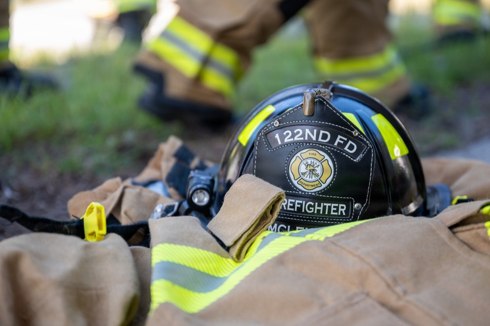 A black firefighter helmet labeled '122ND FD' and 'FIREFIGHTER' with the name 'MCL...' is resting on top of tan turnout gear with yellow reflective striping. The gear appears to be laid out on the ground, possibly during a training exercise or scene response. In the blurred background, at least one firefighter in full turnout gear is visible standing nearby.