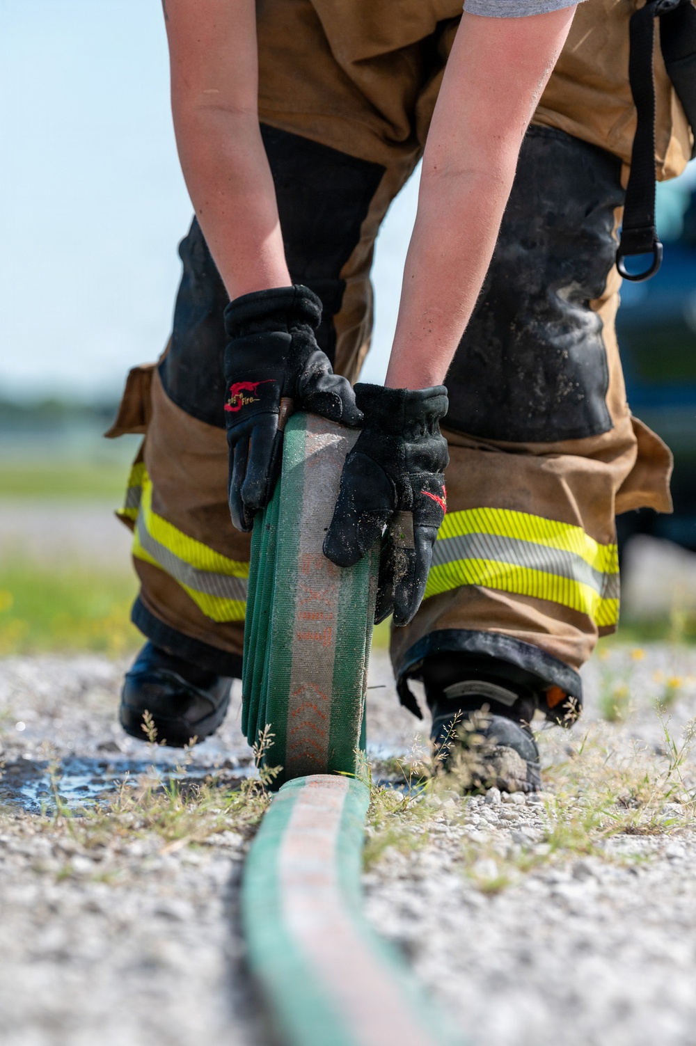 A firefighter in full turnout gear is bending down to handle a flat, deflated fire hose on a gravel surface. The individual is wearing standard structural firefighting protective clothing including tan/brown bunker pants with yellow-green reflective trim stripes and black firefighting gloves. The scene appears to be an outdoor training exercise or post-incident hose management activity.