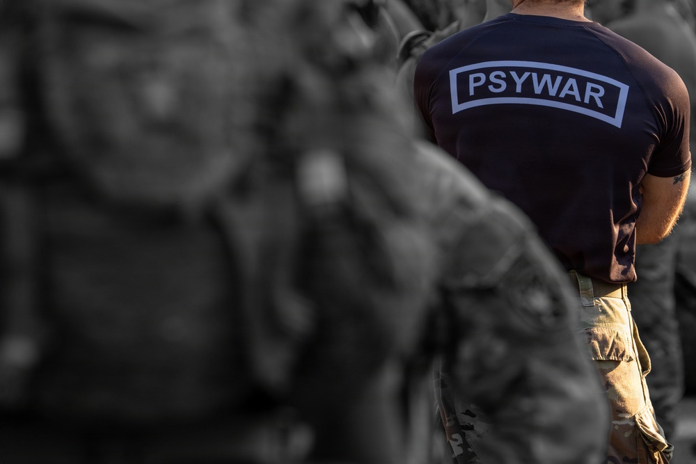 The image shows the back of a person wearing a dark navy blue t-shirt with 'PSYWAR' printed in a rectangular label on the back, paired with tan/multicam military-style cargo pants. The subject appears to be standing among other individuals in military or tactical attire, who are blurred in the background. The selective color treatment keeps the foreground subject in full color while the background figures appear in desaturated tones.