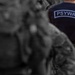The image shows the back of a person wearing a dark navy blue t-shirt with 'PSYWAR' printed in a rectangular label on the back, paired with tan/multicam military-style cargo pants. The subject appears to be standing among other individuals in military or tactical attire, who are blurred in the background. The selective color treatment keeps the foreground subject in full color while the background figures appear in desaturated tones.